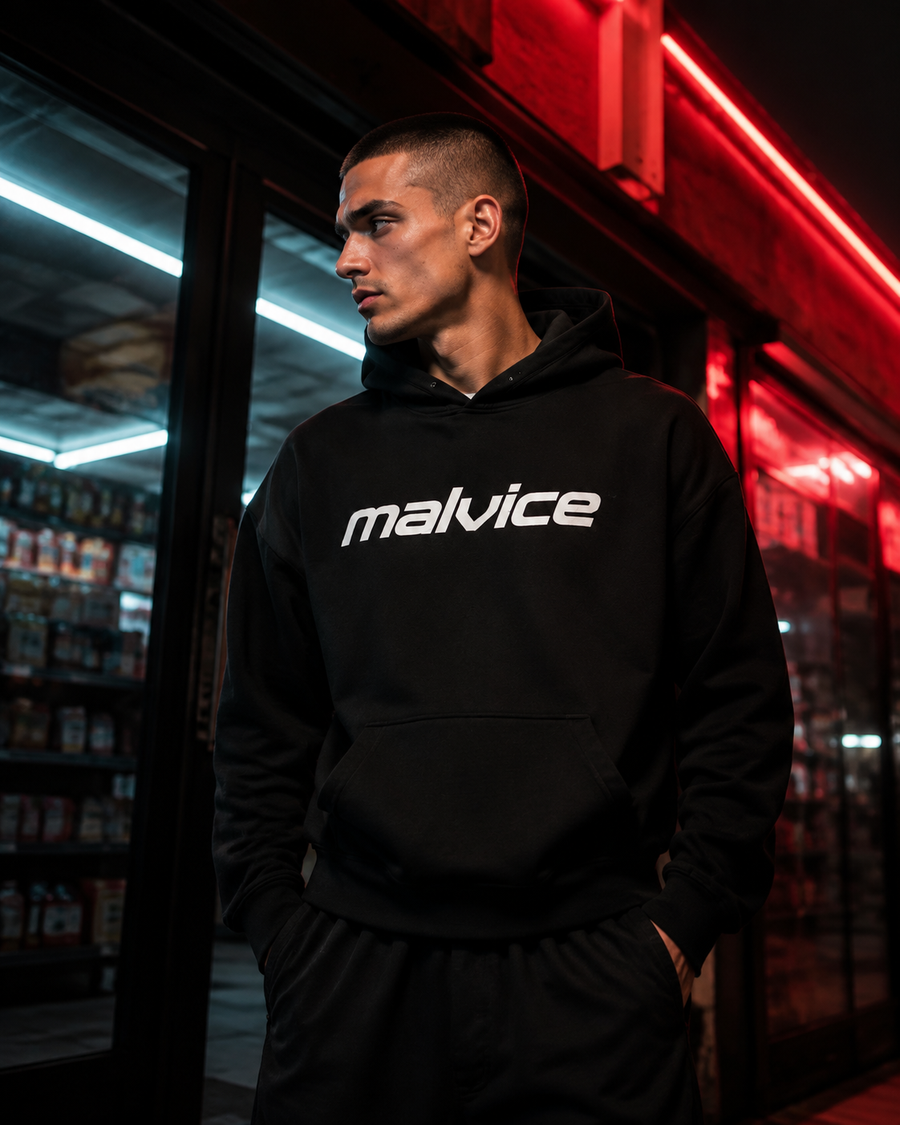 Man wearing a black MALVICE hoodie outside a neon-lit shop