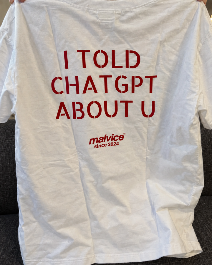 White MALVICE I Told ChatGPT About You oversized T-shirt held indoors