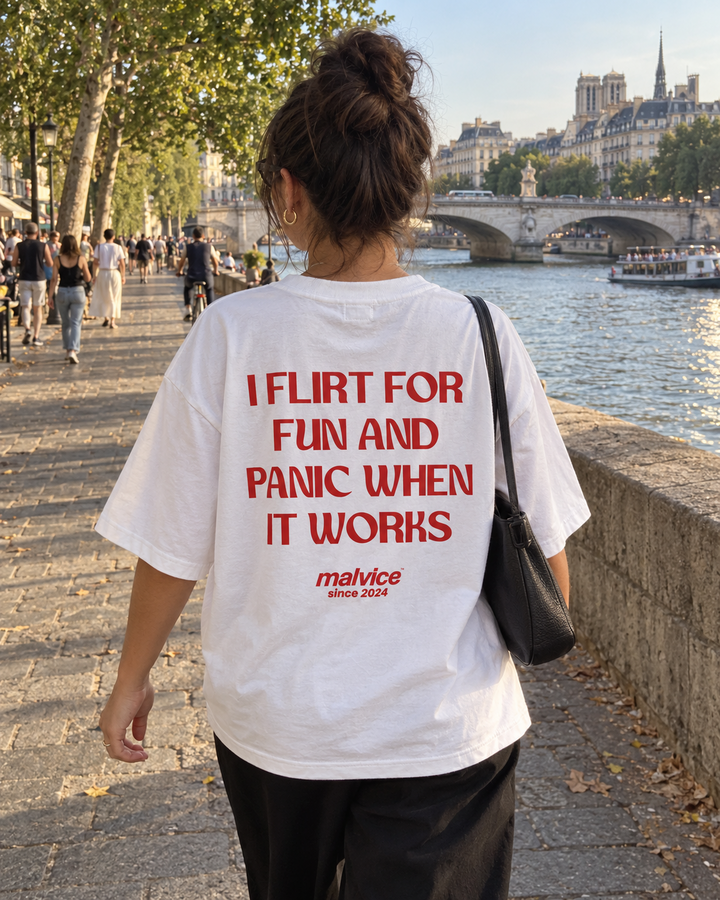 Woman wearing a white MALVICE I Flirt for Fun and Panic When It Works oversized T-shirt beside a city river